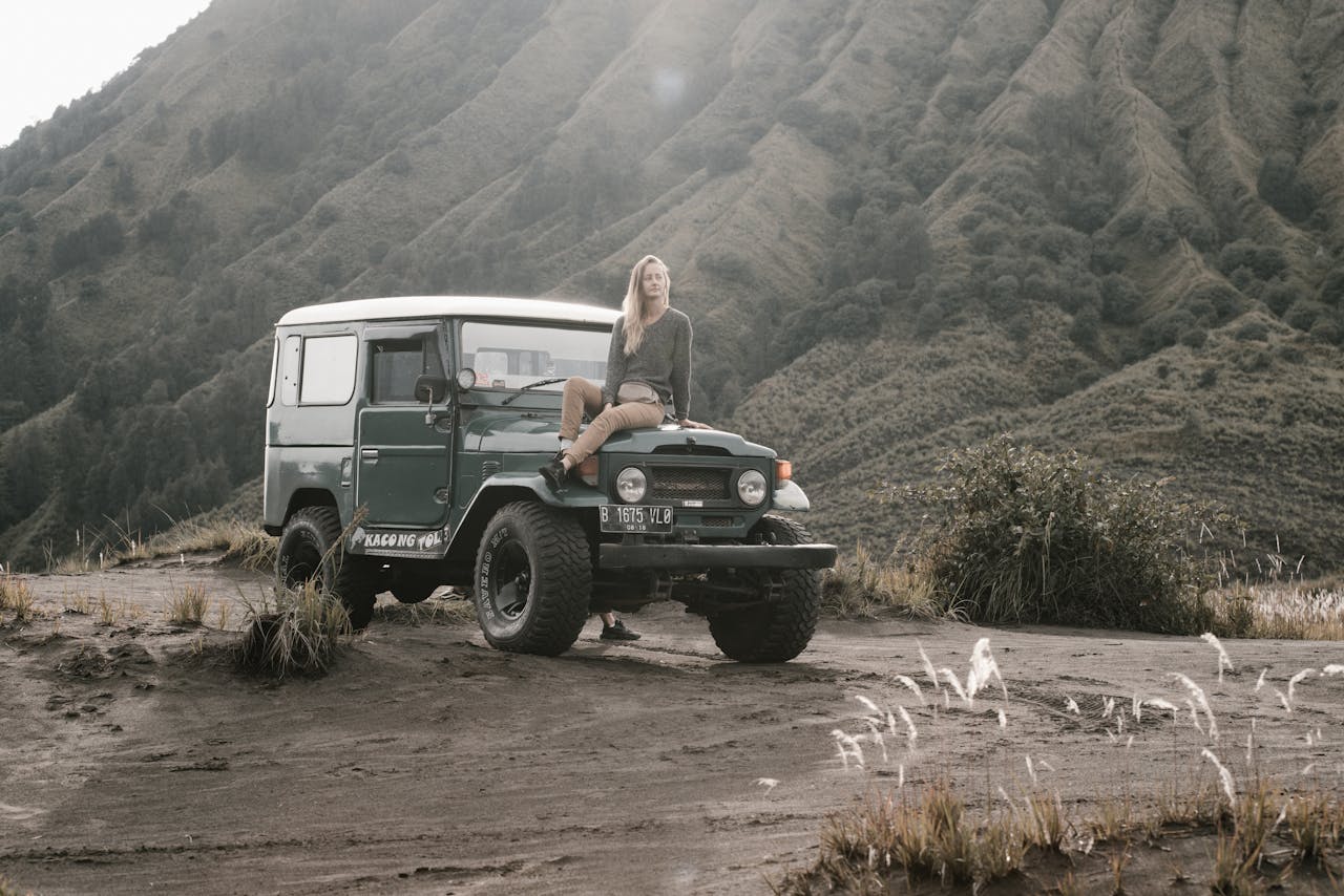 services-01 A woman sits on a jeep in a rugged mountain landscape.