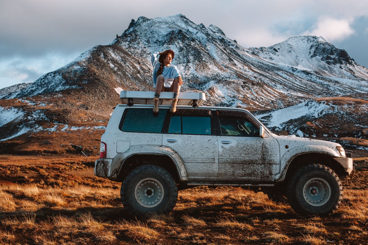 hero-img-01 Full body woman in casual clothes and hat sitting on offroader rooftop against snowy mountain range in sunny cloudy day